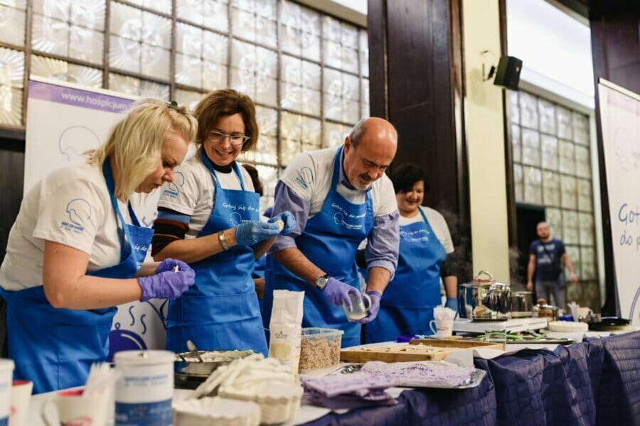 Four people in blue aprons and gloves prepare food at a table filled with cooking supplies, smiling and working together in a bright indoor setting—captured by fotograf eventowy Warszawa.