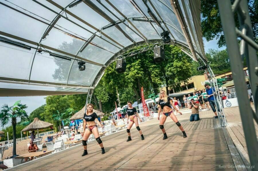 Three women in matching black outfits and knee pads dance on an outdoor stage under a canopy, while people relax on lounge chairs in the sun. Photo coverage from the event captures the vibrant energy of this sunny day.