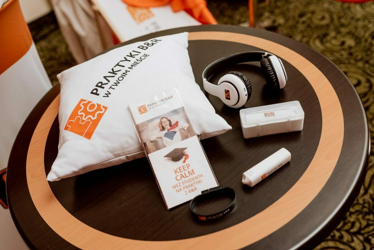 The round table displays an assortment of items, including a pillow with branding, a set of headphones, an informational brochure, a tote and a tube. Both the pillow and brochure have orange and white designs with text. This lively scene captures the essence of event photography against a decorative dark carpet.  