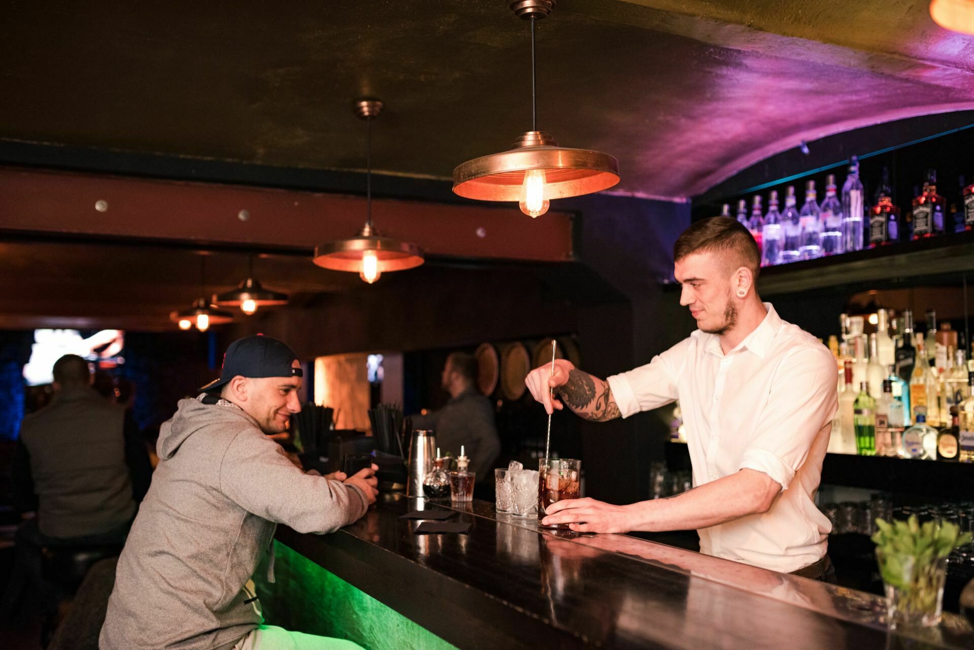 A bartender in a white shirt mixes a drink for a man wearing a baseball cap