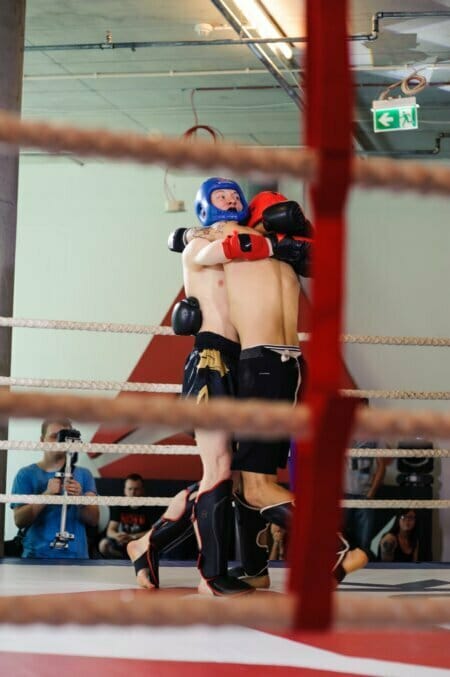 Two fighters, one in blue and the other in red headgear, wrestle in a boxing ring. They wear shin guards and gloves. Spectators and a photographer recording the event can be seen outside the ring, with the ropes partially obscuring the view. Above, an exit sign is visible.   