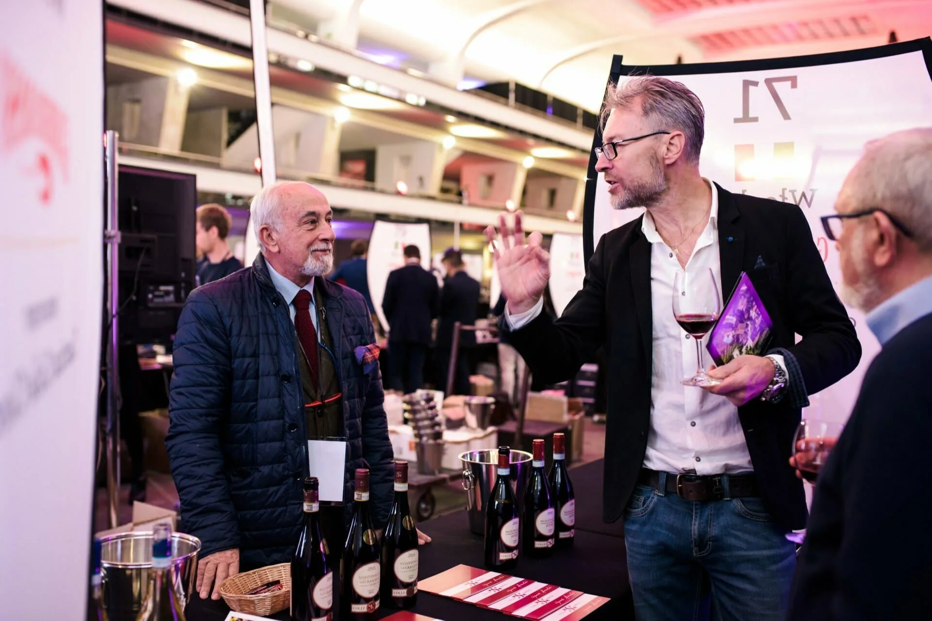 Three men enjoy a wine-tasting event, with one enthusiastically sharing his thoughts while holding a glass of red wine. Bottles and tasting materials are displayed on the table—a perfect scene for fotografia eventowa Warszawa.