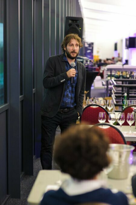 A man in a jacket and plaid shirt with long hair holds a microphone while standing next to a table full of wine glasses