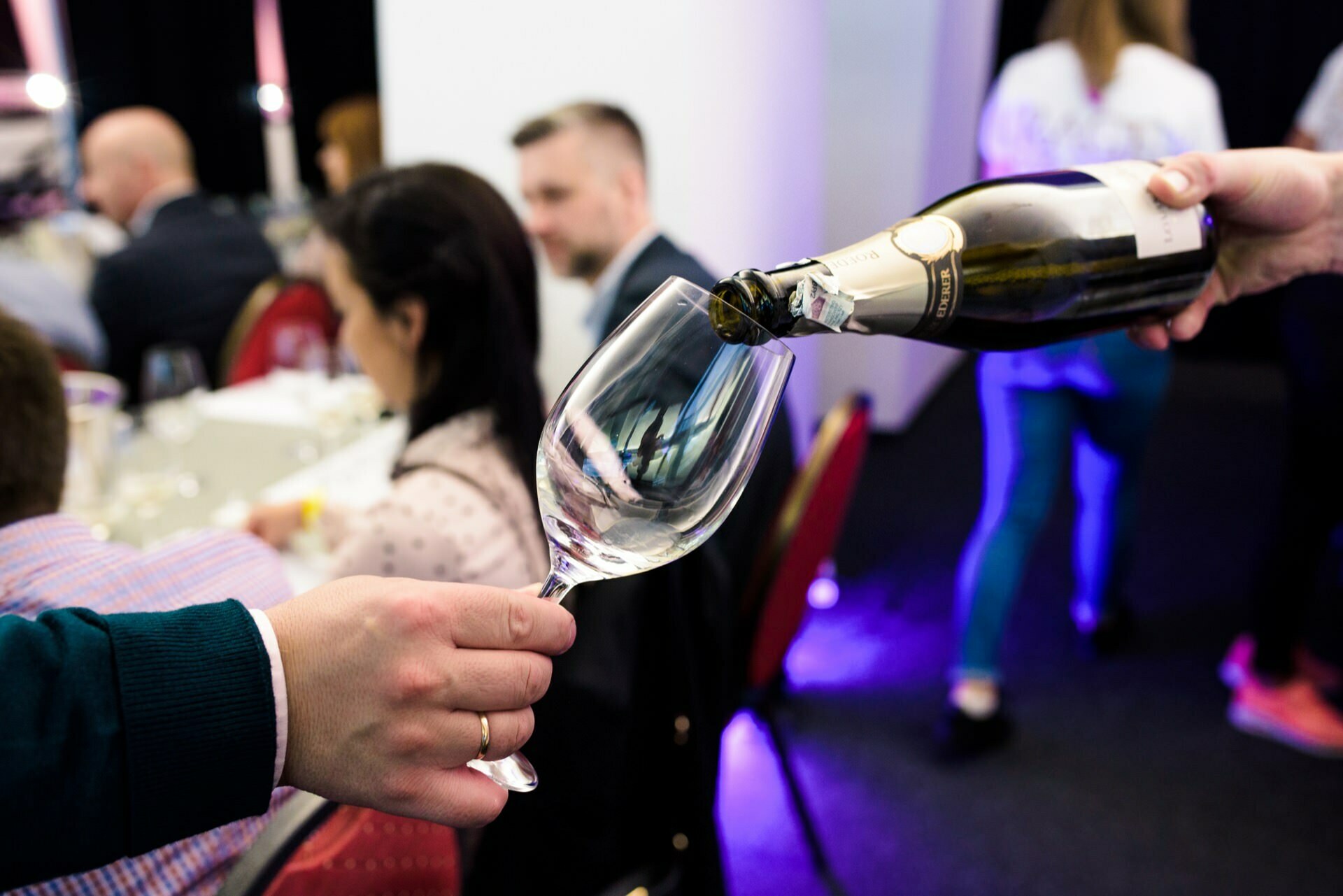 Close-up of a glass that a man's hand with a ring is holding and someone pours wine from a bottle into that glass