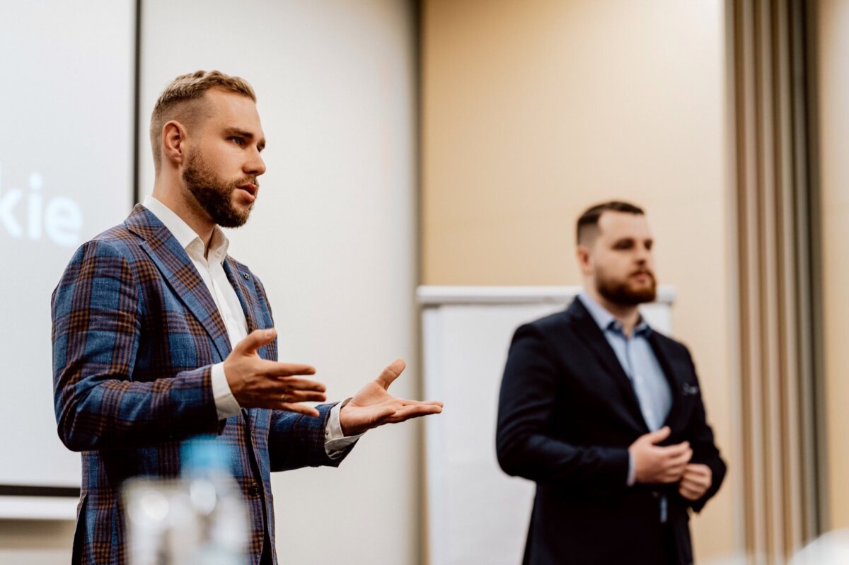 Two men in business attire appear in the conference room. The man on the left, in a blue plaid suit, gestures with his hands as he speaks. The man on the right, in a dark suit, stands attentively. A projector screen and blackboard are visible in the background; this captures a dynamic photo-report of the events.   