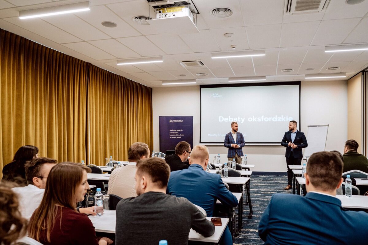 Grupa osób siedzi przy stołach w sali konferencyjnej z żółtymi zasłonami, naprzeciwko ekranu projektora, na którym widnieje napis „Debaty oksfordzkie”. Z przodu sali stoi dwóch mężczyzn, przemawiających do publiczności. Fotorelacja z wydarzenia Marcina Krokowskiego uwiecznia każdą chwilę za pomocą flipchartu umieszczonego z boku.