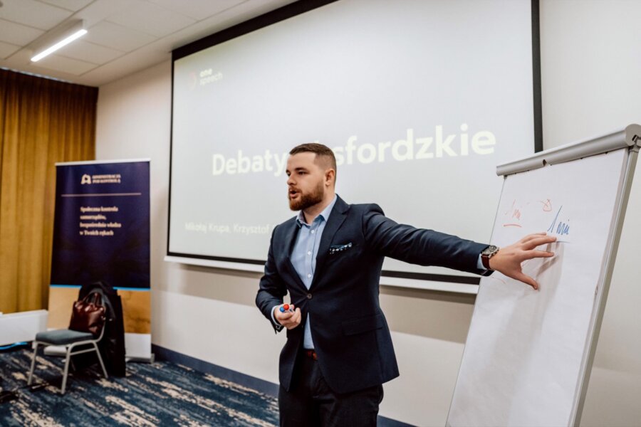 Mężczyzna w garniturze wskazuje na flipchart w sali konferencyjnej. Na ekranie za nim widnieje napis „Debaty Oksfordzkie”. Trzyma marker i wydaje się być zaangażowany w prezentację. W tle baner i krzesła, pięknie uchwycone przez Marcina Krokowskiego, doświadczonego fotografa eventowego Warszawa.