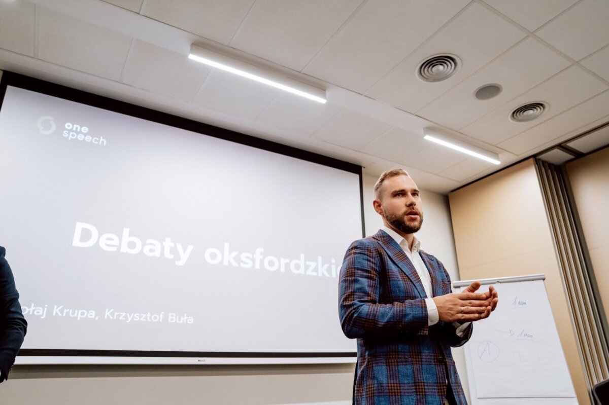 W sali konferencyjnej przemawia mężczyzna w kraciastym garniturze, stojący przy flipcharcie. Za jego plecami na dużym ekranie wyświetlany jest polski tekst „Debaty oksfordzkie” oraz logotypy, w tym „jedno przemówienie”. Powyżej widoczne są świetlówki i płytki sufitowe, które oddają atmosferę wydarzenia w tej fotorelacji z eventu.