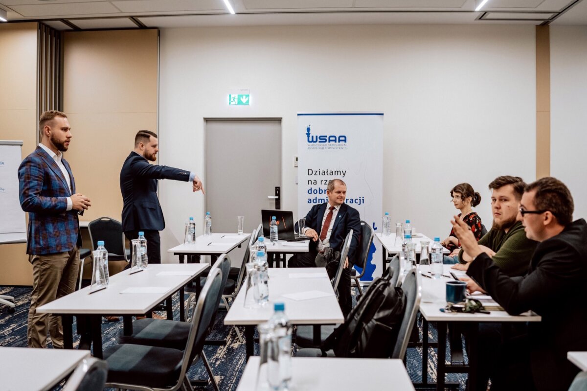 Grupa osób w sali konferencyjnej zaangażowana w dyskusję. Dwóch mężczyzn stoi, jeden skierowany w bok, pozostali siedzą przy stołach z notatnikami i butelkami z wodą. W tle widoczny baner z napisem „WSAA”, pięknie uchwycony przez Marcina Krokowskiego, zdolnego fotografa eventowego.