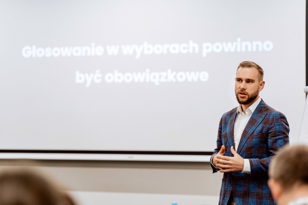 Mężczyzna w kraciastym garniturze przemawia przed ekranem projekcyjnym, na którym widnieje polski napis „Głosowanie w wyborach powinno być obowiązkowe”. Wygląda na to, że wygłasza prezentację lub wykład. Mogłaby z tego powstać ciekawa fotorelacja z wydarzeń autorstwa fotografa eventowego.