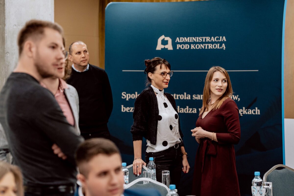 Na imprezie zbiera się grupa ludzi. Na pierwszym planie widoczne są dwie kobiety i trzech mężczyzn. Za nimi znajduje się niebieski baner z białym tekstem i logo u góry z napisem „ADMINISTRACJA POD KONTROLĄ”. Na stołach ustawiane są butelki z wodą, utrwalające chwile na potrzeby fotografii eventowej Marcina Krokowskiego.