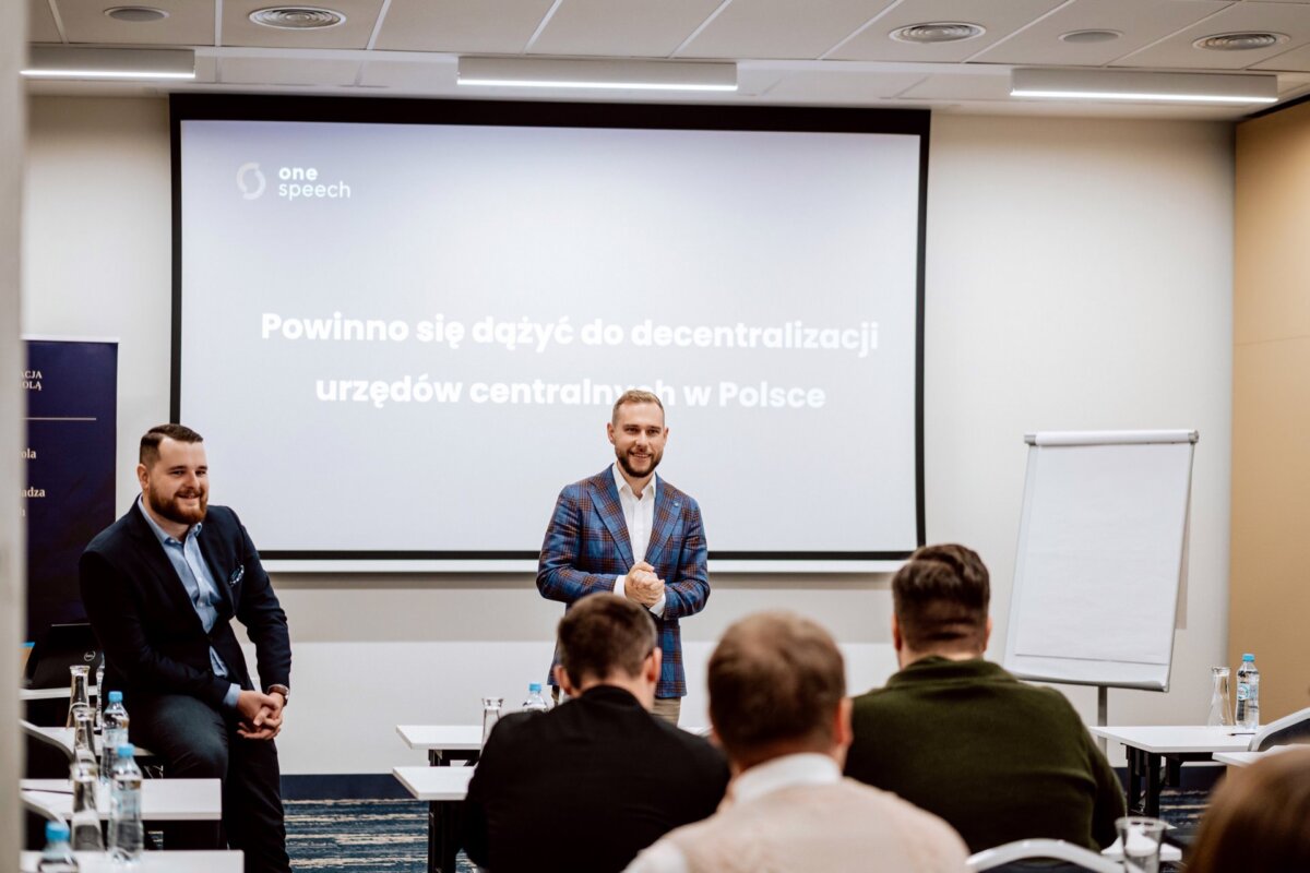 Mówca stoi przed wyświetlanym slajdem w sali konferencyjnej i przemawia do publiczności siedzącej przy stołach. Na slajdzie widnieje napis: „Powinno podążać do decentralizacji urzędów centralnych w Polsce”, co oznacza „Należy podjąć wysiłki na rzecz decentralizacji urzędów centralnych w Polsce”. Ujęcie autorstwa fotografa eventowego Warszawa.