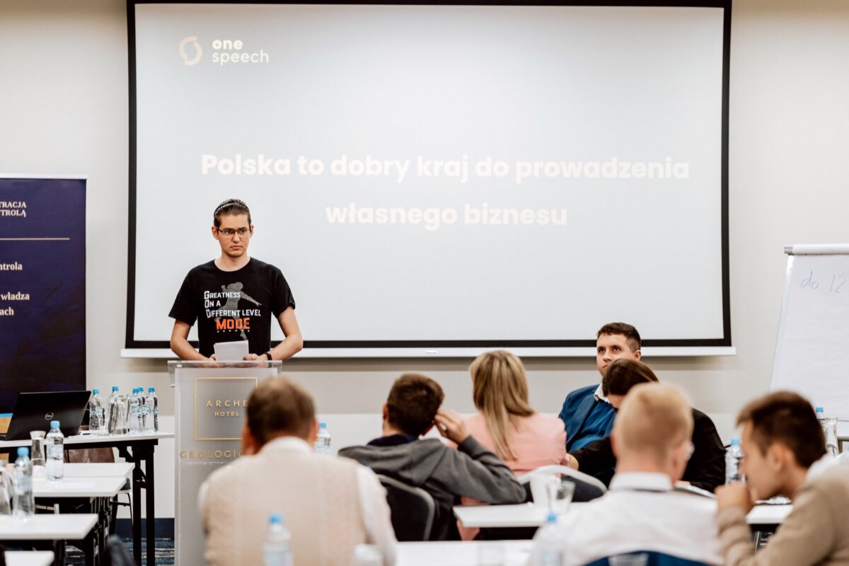 Mężczyzna stoi na podium pod logo „jednego przemówienia”, występując przed publicznością w sali konferencyjnej. Z tyłu ekranu widnieje napis „Polska to dobry kraj do prowadzenia własnego biznesu”, co oznacza „Polska to dobry kraj do prowadzenia własnego biznesu”. Ten moment został pięknie uchwycony w fotorelacji z eventu.