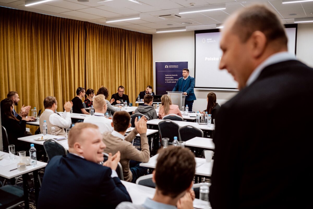 Mężczyzna stoi na podium i przemawia do publiczności w sali konferencyjnej. Uczestnicy siedzą przy stołach, niektórzy klaszczą, inni są pochłonięci rozmową. Za prelegentem widoczny jest duży ekran prezentacyjny i baner. Atmosfera sprawia wrażenie profesjonalnej, uchwyconej żywej fotorelacji z wydarzeń.