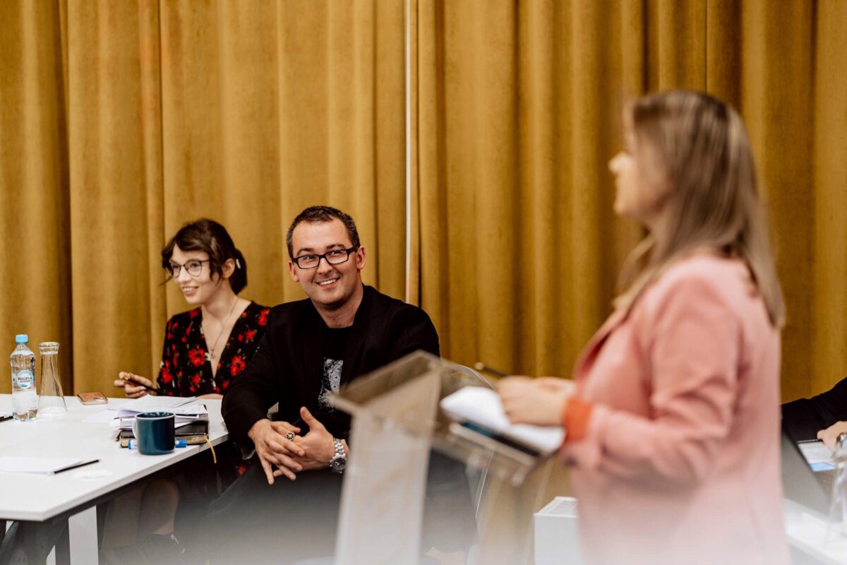 Kobieta w różowej marynarce przemawia na podium w sali konferencyjnej. W tle Marcin Krokowski, fotograf eventowy Warszawa, uśmiecha się, siedząc przy stole z laptopem. Obok niego siedzi inna kobieta w okularach i kwiecistej sukience, skupiona na swoich notatkach.