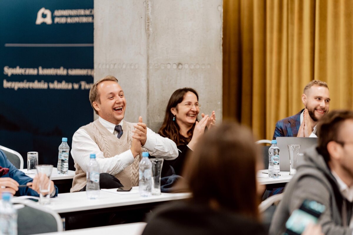 Trzy osoby siedzą przy stole konferencyjnym z notatnikami i butelkami z wodą, klaszcząc i uśmiechając się, co sugeruje, że cieszą się prezentacją. W tle widać zasłony i część szyldu na ścianie, co uchwyca żywą fotorelację z eventu.