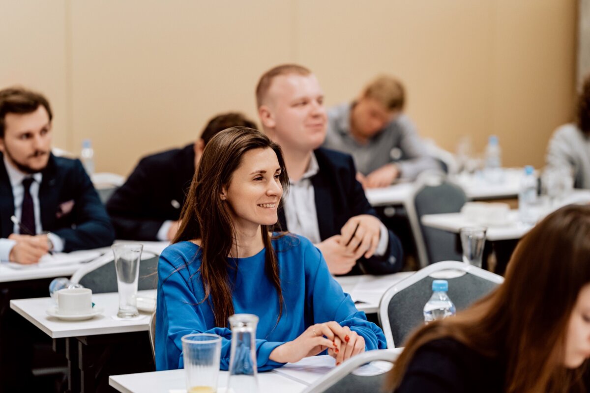 Kobieta w niebieskiej koszuli siedzi i uśmiecha się na konferencji lub seminarium, w tle kilku innych uczestników. Uczestnicy siedzą przy stołach, przed którymi stoją szklanki, kubki i dokumenty. Oddaje to istotę wydarzenia autorstwa Marcina Krokowskiego, znanego fotografa eventowego Warszawa.