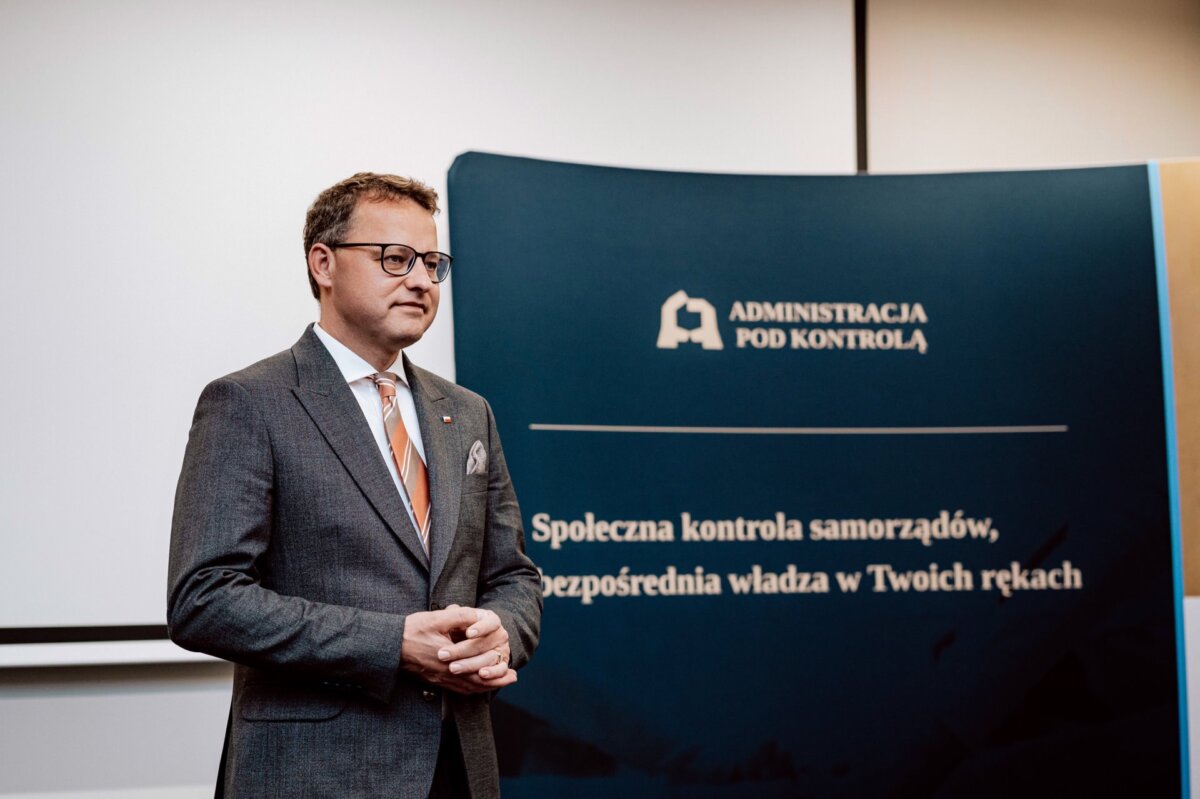 A man in a gray suit and light brown tie stands in front of a blue banner with white text, presumably at an official event taking place indoors. The banner reads "ADMINISTRATION UNDER CONTROL" and contains additional text in Polish. It could be part of a photo essay of the event or captured by an event photographer.  