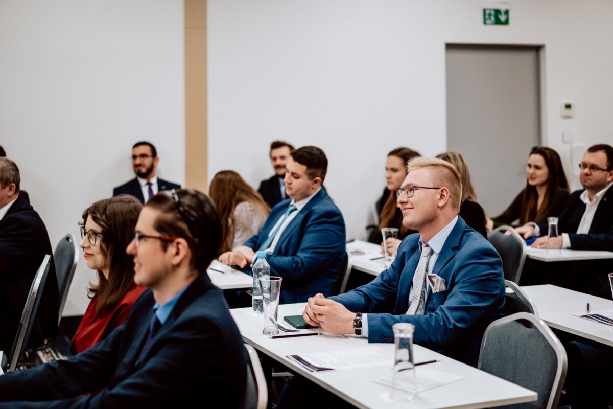 Grupa osób, głównie w strojach biznesowych, siedzi uważnie w sali konferencyjnej. Niektórzy robią notatki, inni słuchają. Uczestnicy siedzą przy stołach, a wśród nich jest mieszanka mężczyzn i kobiet, wszyscy skupieni w stronę niewidzianej prezentacji lub mówcy. Fotografia eventowa doskonale utrwala takie momenty.