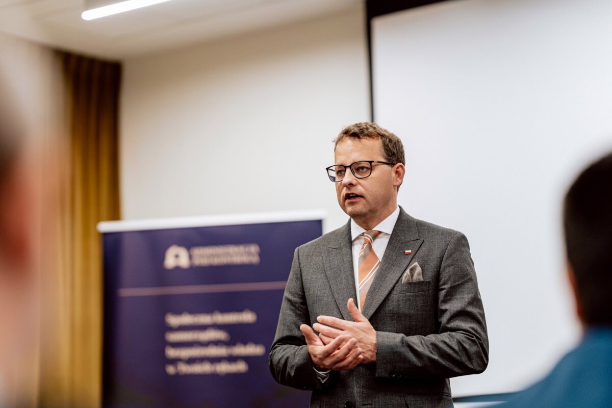 Przed publicznością przemawia mężczyzna ubrany w garnitur i krawat. Za nim znajduje się stojący baner z częściowo zasłoniętym tekstem i dużym, pustym ekranem projekcyjnym. Sceneria wydaje się mieć charakter formalny, być może konferencja lub wykład, uchwycił Marcin Krokowski, znany fotograf eventowy Warszawa.