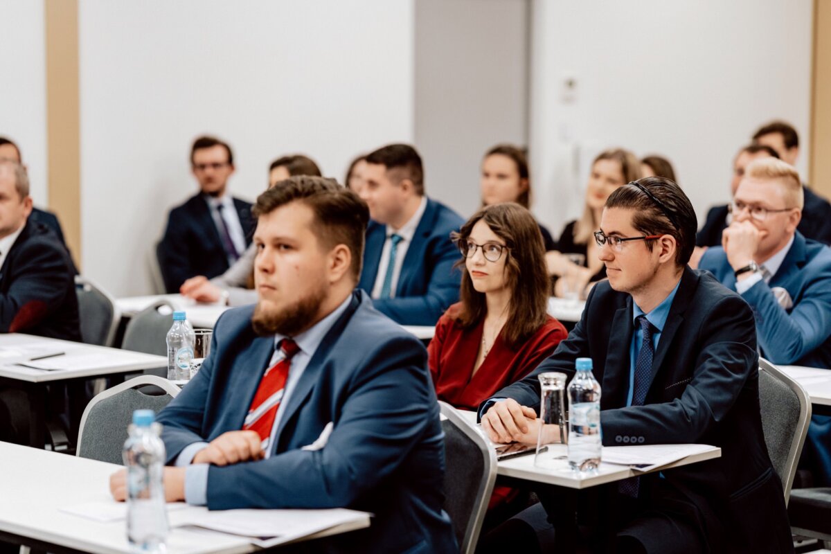 Grupa osób ubranych w profesjonalny strój siedzi uważnie w sali konferencyjnej, zwrócona twarzą do przodu. Niektórzy mają notesy i długopisy na stołach przed sobą, a na każdym biurku stoi woda butelkowana. Sala utrzymana jest w biało-beżowej kolorystyce, idealnej do fotografii eventowej.