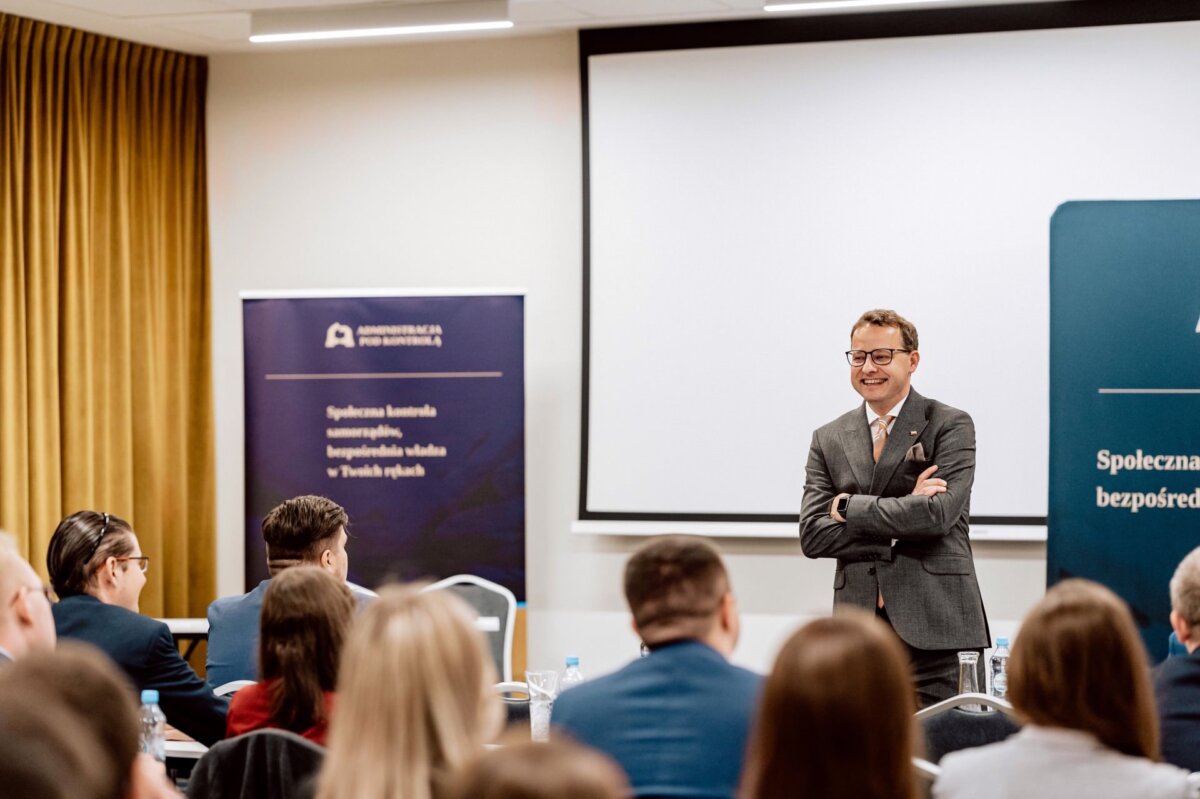 Mężczyzna w garniturze stoi uśmiechnięty i przemawia przed ekranem projektora w sali konferencyjnej wypełnionej siedzącymi uczestnikami. Po jego prawej stronie wyświetlany jest baner z tekstem i logo, a w tle widoczna jest złota kurtyna, która uchwyciła istotę profesjonalnej fotografii eventowej.