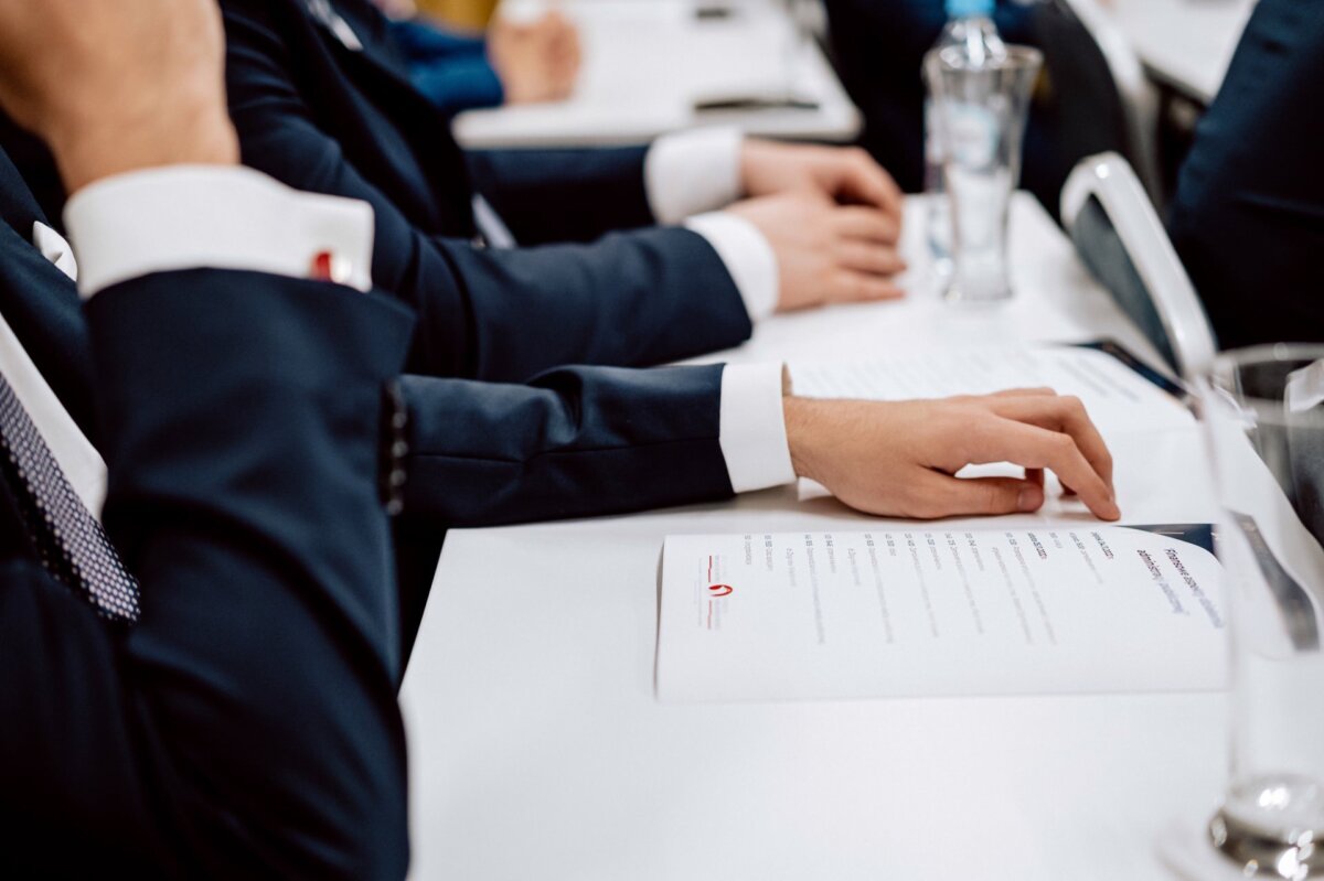 Zbliżenie kilku osób w formalnych garniturach siedzących przy stole konferencyjnym lub na spotkaniu. Niektórzy trzymają długopisy, a na stole przed nimi leży dokument lub agenda. W tle widoczne są również butelki wody i szklanki, uchwycone przez znanego fotografa eventowego Marcina Krokowskiego.