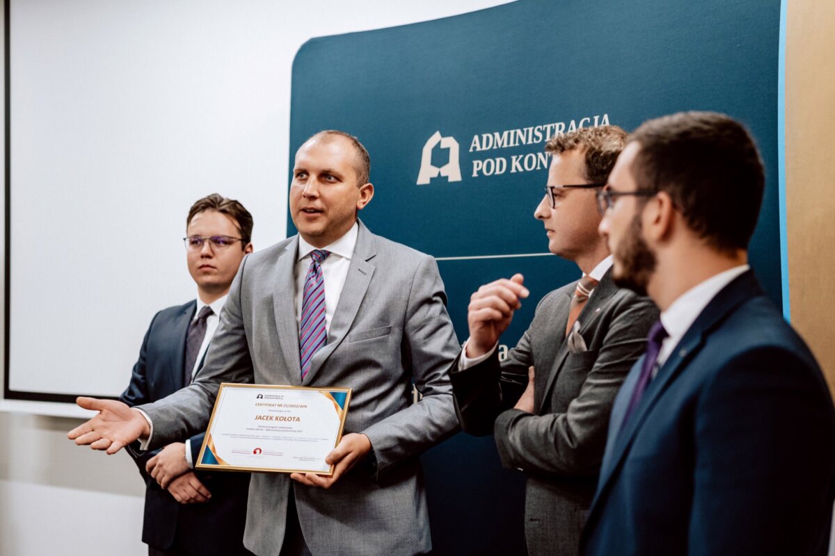 Czterech mężczyzn w strojach biznesowych stoi na tle napisu „Administracja Pod Kontrolą”. Mężczyzna drugi od lewej trzyma certyfikat. Sprawiają wrażenie pochłoniętych rozmową lub prezentacją, idealną do fotorelacji z wydarzeń przez każdego doświadczonego fotografa eventowego warszawa.