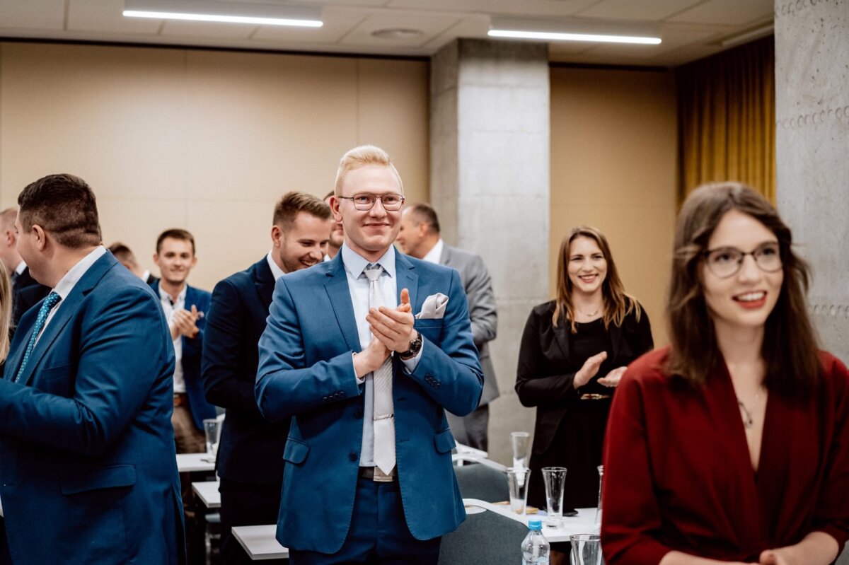 Grupa osób w strojach biznesowych stoi i klaszcze w czymś, co przypomina salę konferencyjną. Mężczyzna na pierwszym planie ma na sobie niebieski garnitur i okulary, podczas gdy w pobliżu uśmiecha się kobieta w czerwonej marynarce. Ta fotorelacja z wydarzeń pokazuje, jak wszyscy klaszczą i uśmiechają się, co świadczy o powodzeniu wydarzenia.