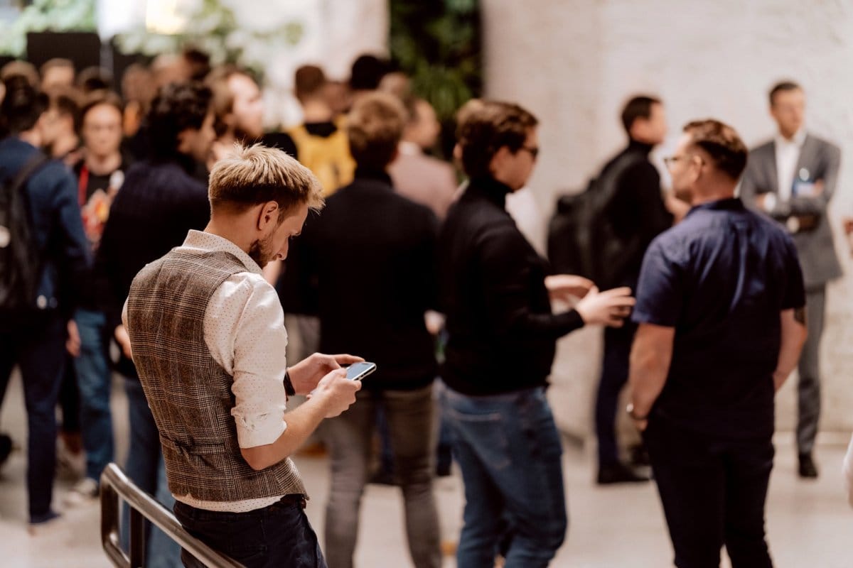 A man in a vest and white shirt leaning against a railing against a background of a group of people having a conversation