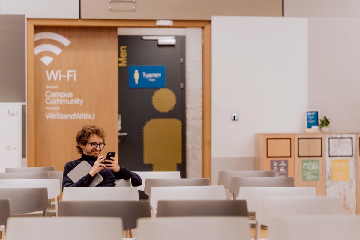 A boy with glasses sits alone on one of the white chairs and smiles into the phone