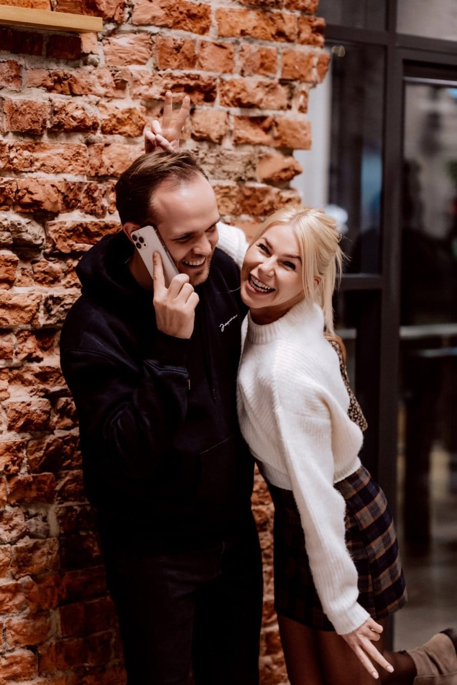 Boy talks on phone and blonde in white sweater hugs him against brick wall