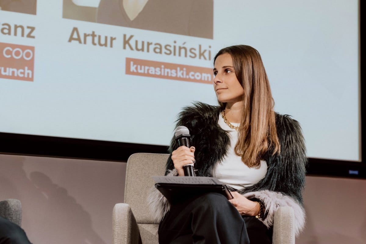 Girl in white blouse and fur jacket sits with microphone holding documents