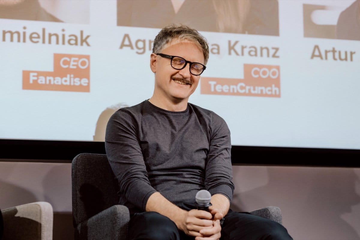 A man with a mustache and glasses holds a microphone and laughs with his eyes closed