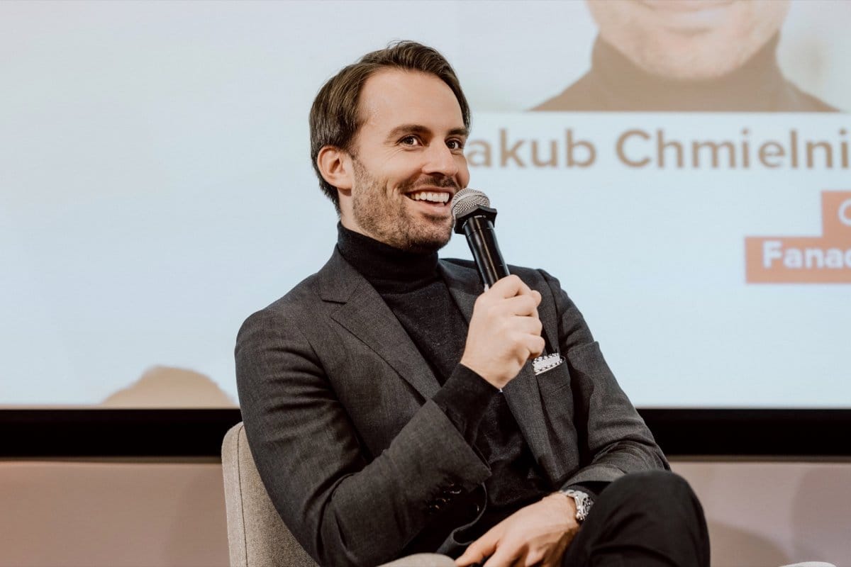 A man in a gray jacket and black turtleneck smiles and speaks into the microphone