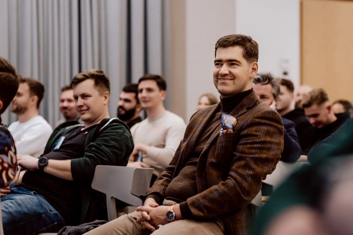 A man in a brown jacket smiles while sitting in the audience