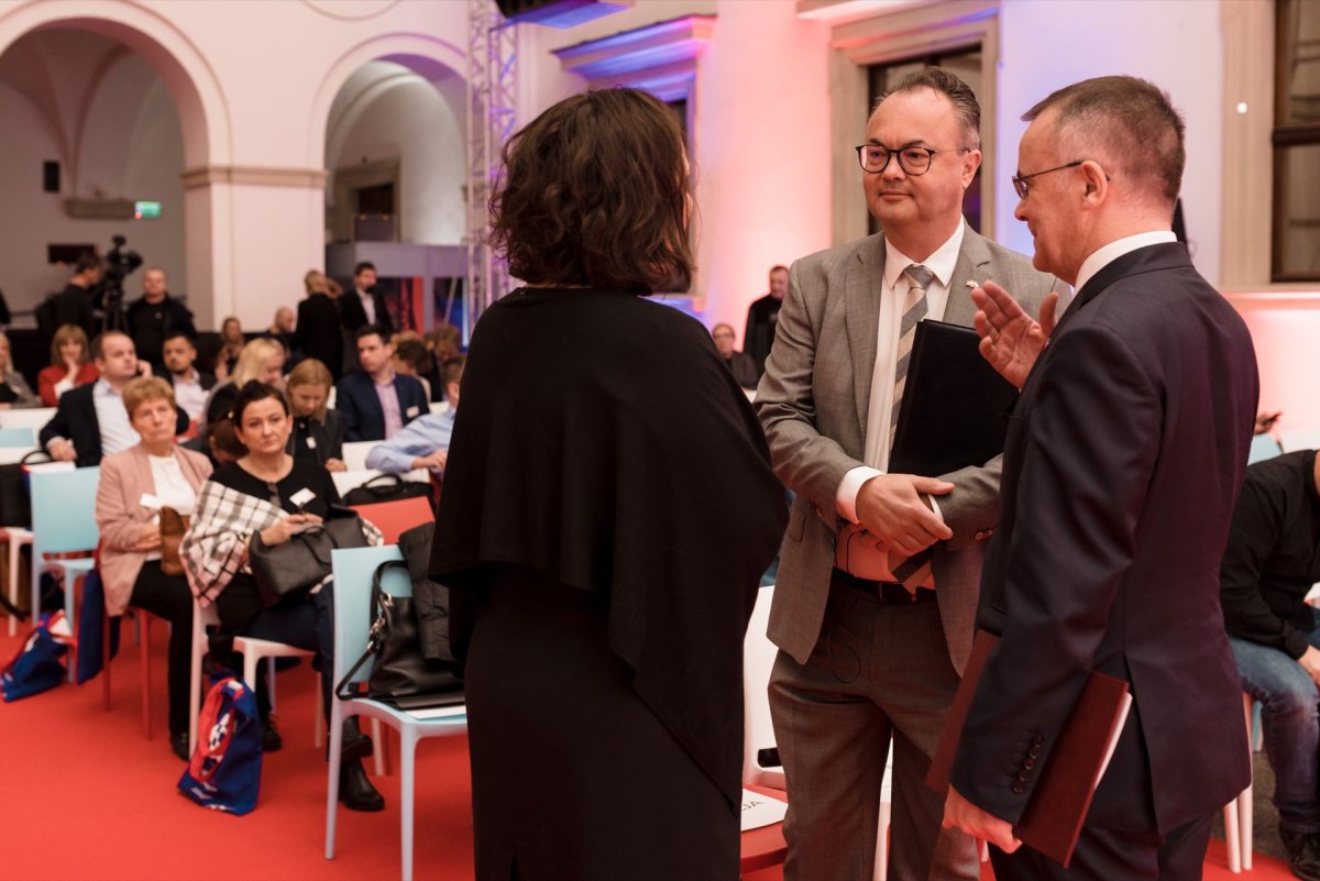 A group of people in a formal setting, with two men and one woman standing and talking in the foreground. The men are dressed in suits, and the woman is standing with her back to the camera. In the background are seated people who appear to be involved in the event - an encouraging photo-op.  