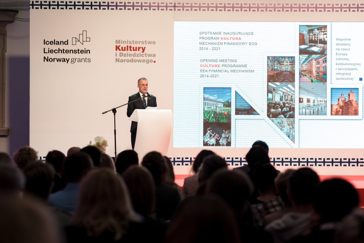 A man in a suit stands at the podium and addresses the audience. Behind him is a large screen displaying text and images related to the "Opening Meeting Culture Program" for grants from Iceland, Liechtenstein and Norway. The room is dimly lit with focused light on the speaker, captured by marcin krokowski, an event photographer.  