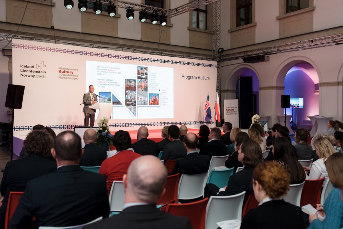 The speaker stands at a podium, addressing a seated audience in a modern conference room. Behind him, a presentation slide titled "Culture Program" with images and text is displayed on a large screen. National flags have been placed on the stage next to the speaker, capturing the essence of the event in a photo essay of the event.  