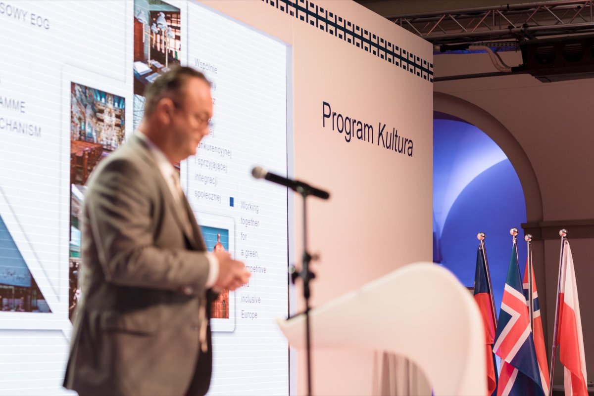 A person in a suit stands at a podium with a microphone, presenting at a conference or event. A large screen displays slides with the words "Culture Program." Several flags can be seen in the background, suggesting an international context. This scene can be perfectly captured for event photo coverage.   