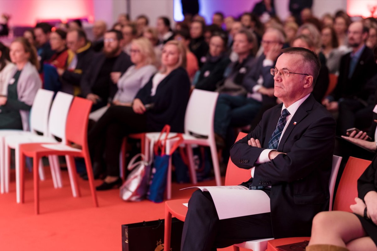 A man in a suit and tie sits with his arms crossed in a red chair in a large conference room and appears focused. Behind him is an audience seated in red and white chairs. The room is lit up in shades of pink and purple, capturing the essence of this photo event.  