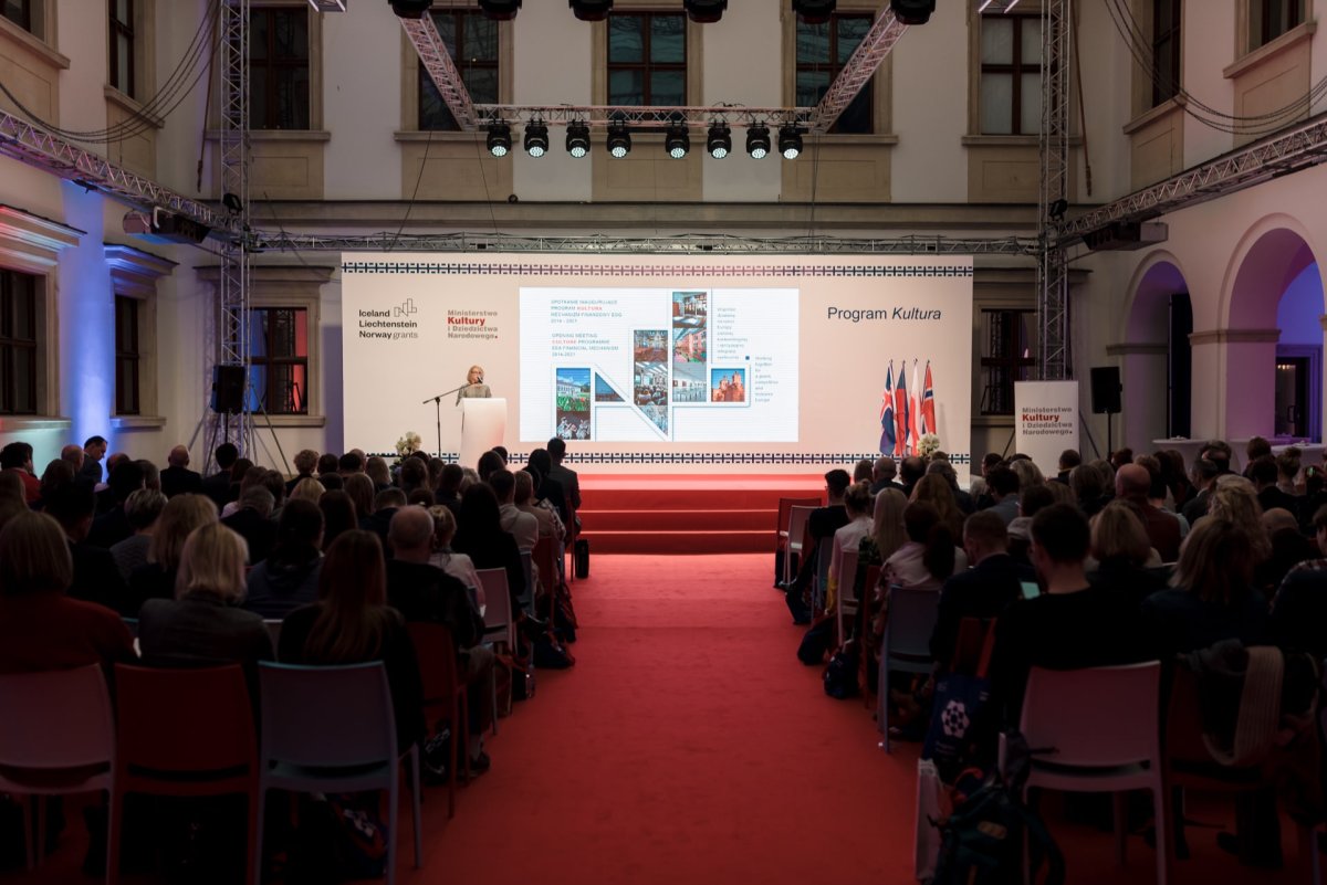 The presenter stands on a lighted stage and delivers a presentation to an audience in a large room with a red carpet. A projection screen at the back displays informational slides. On the right side of the stage, national flags are flown, and the room is filled with rows of chairs displaying a photo-op of Marcin Krokowski's event.  