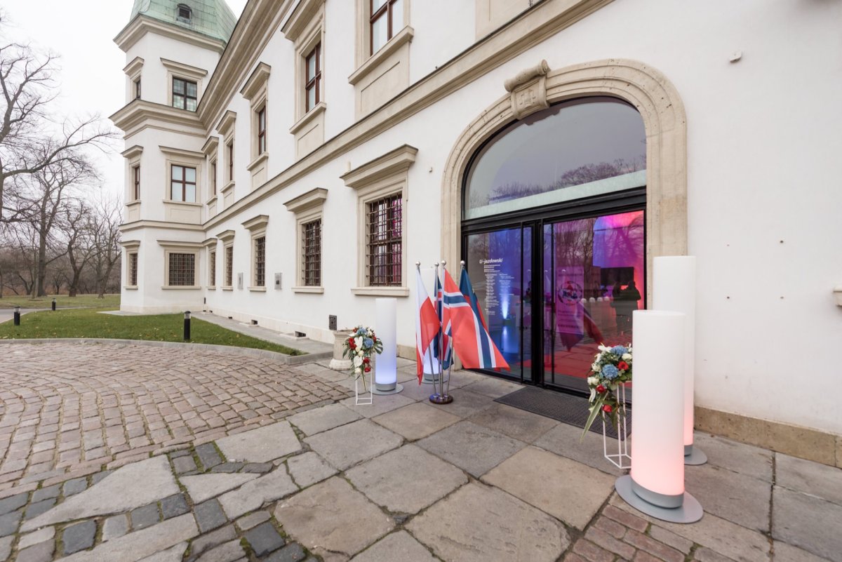 This photo shows the entrance to an elegant building with large arched double glass doors. Two small podiums with flower arrangements flank the entrance along with two flagpoles with red, white and blue flags. The building's classic white facade is beautifully captured, making it ideal for event photo coverage.  