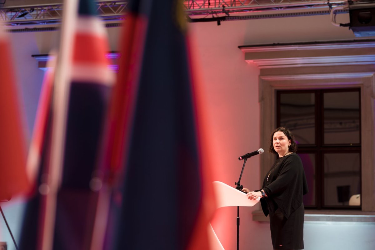 A woman stands on a podium with a microphone and looks as if she is speaking or preparing to speak. In the background is a high ceiling with exposed metal beams and a large window. In the foreground, blurry national flags are visible - the perfect moment for an event photo essay captured by an experienced event photographer. Warm lighting adds to the atmosphere.   