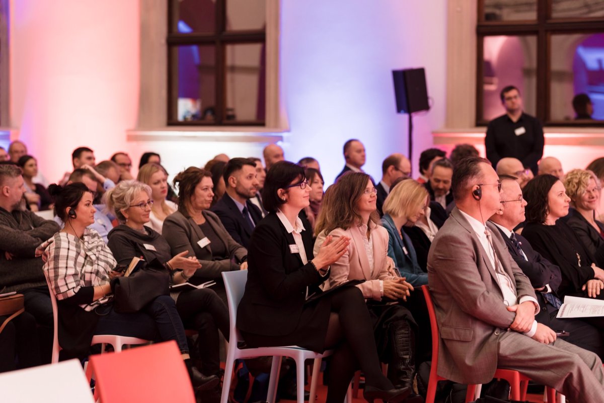 A diverse group of people dressed in business attire sit in rows of chairs at the conference. Some attendees clap, others listen intently. The room is softly lit, with a mix of shades of purple and pink, perfectly captured by event photography, highlighting every detail.  