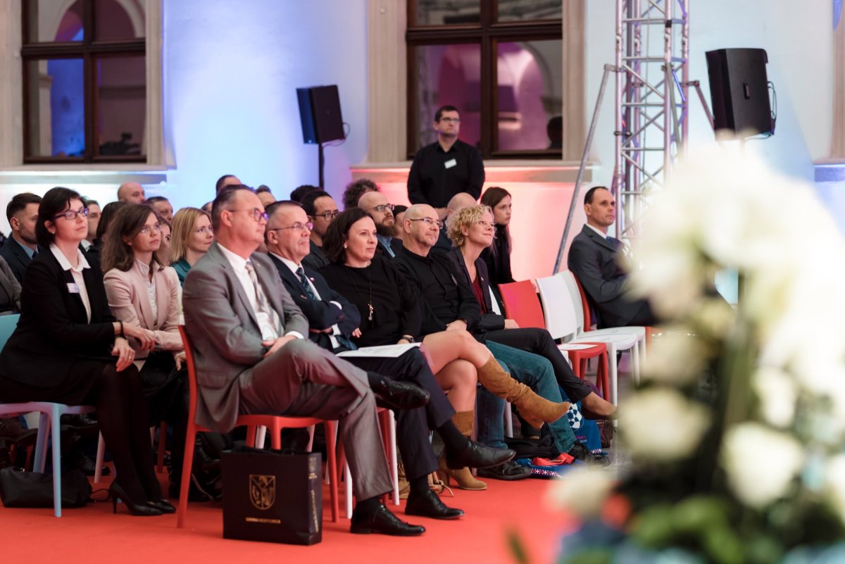 A group of people dressed in business and casual attire sit and listen attentively to a presentation in a warmly lit room with a red carpet. In the foreground is a blurred floral composition. Marcin Krokowski captured this moment as part of his photo report of the events.  