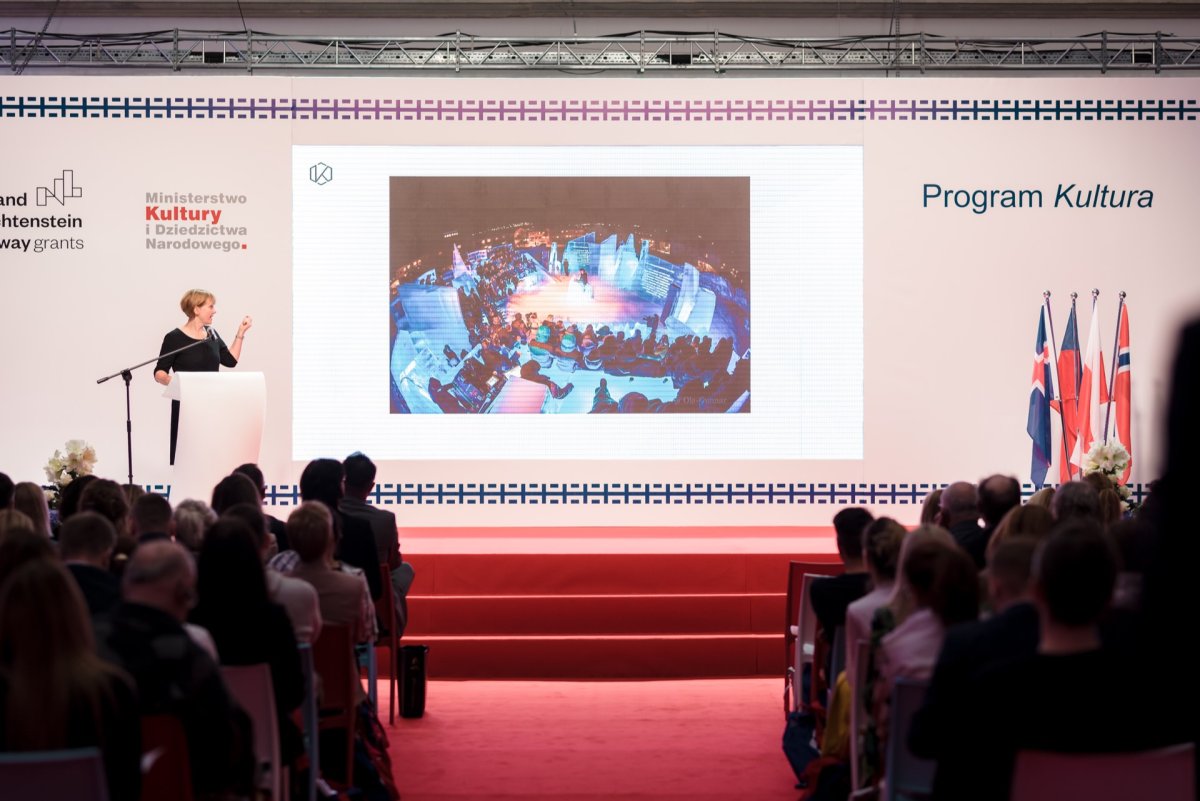 A woman stands at a podium on a stage covered with a red carpet and performs before an audience. Behind her back, a large screen projects an image of the circular hall. Next to the screen are the logos of grants and ministries, while on the right side of the stage are several flags that capture the essence of the photo-report of the events.  