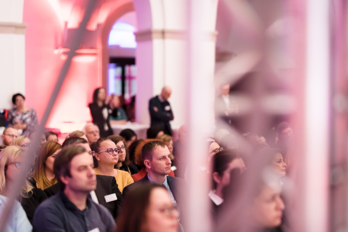 A diverse group of people sit in a room with white arches and pink lighting and listen attentively to Marcin Krokowski. Some people stand in the back. The atmosphere is formal, suggesting a photo essay of an event like a conference or seminar.  