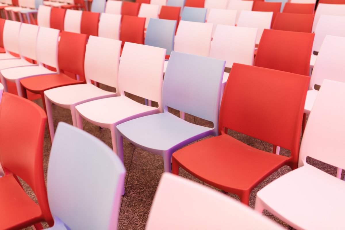 A carefully arranged collection of red, white and one blue chair occupies the auditorium, creating a visually striking pattern. The chairs are made of plastic and are all empty and waiting for attendees. The floor is covered with a mottled gray carpet. Captured by Marcin Krokowski, event photographer warszawa.   
