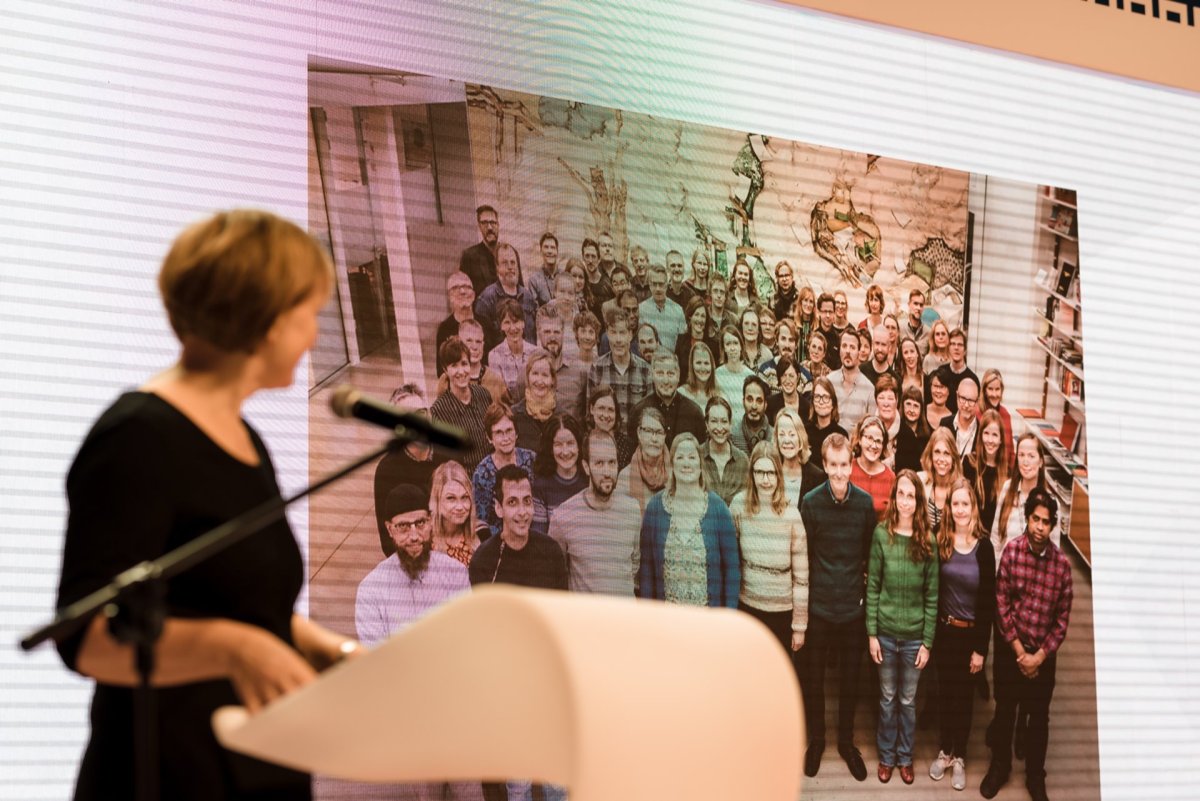 A woman speaks at the podium in concentration, while a large screen behind her displays a group photo of more than fifty people standing close together and smiling. In the background of the screen, a map of the world can be seen, indicating the diverse international setting - a perfect photo-op of the events. 